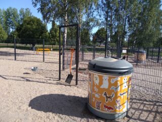 Peltosaaren koirapuiston aitaukset, etualalla iso roskis, jonka kyljessä piirroskuvia koirista.