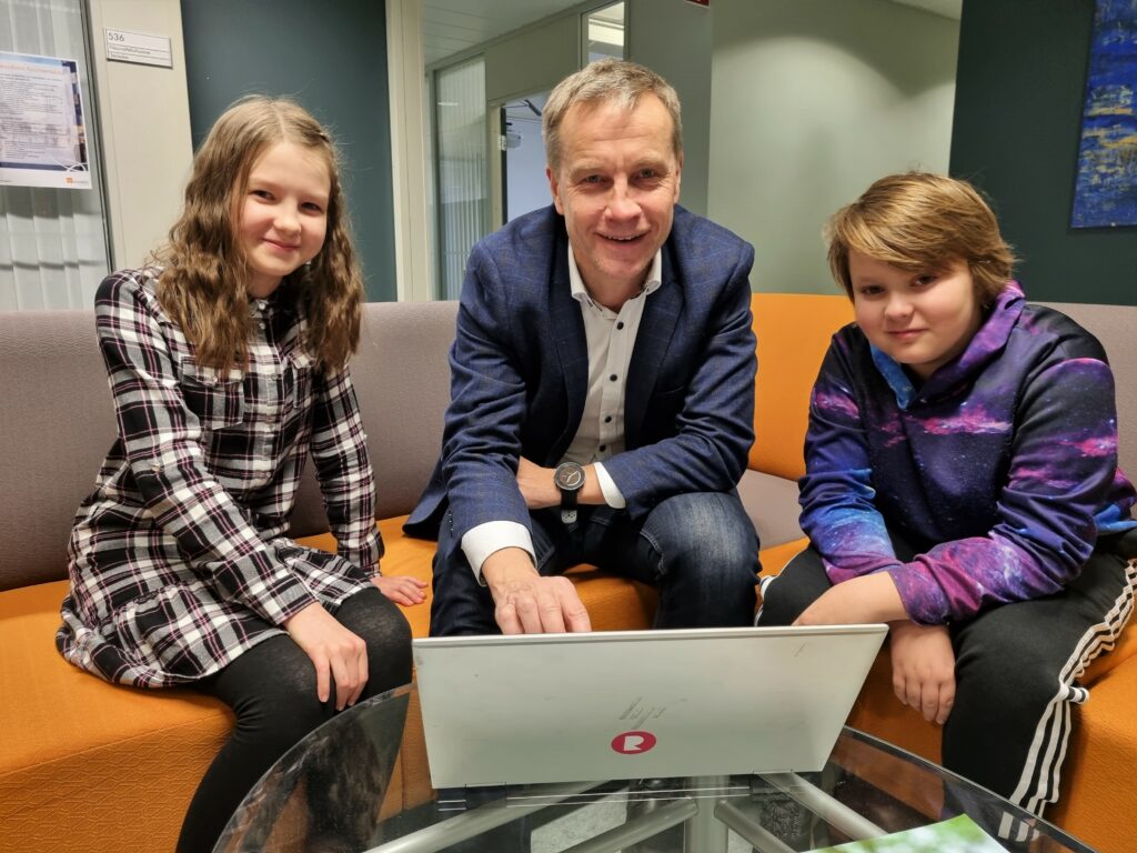 Viidesluokkalainen tyttö, Sofia Pesonen, kaupunginjohtaja Jere Penttilä sekä toinen viidesluokkalainen Santeri Hakala istuvat keltaisella sohvalla. Edessään heillä on pöydällä avoinna oleva kannettava tietokone.