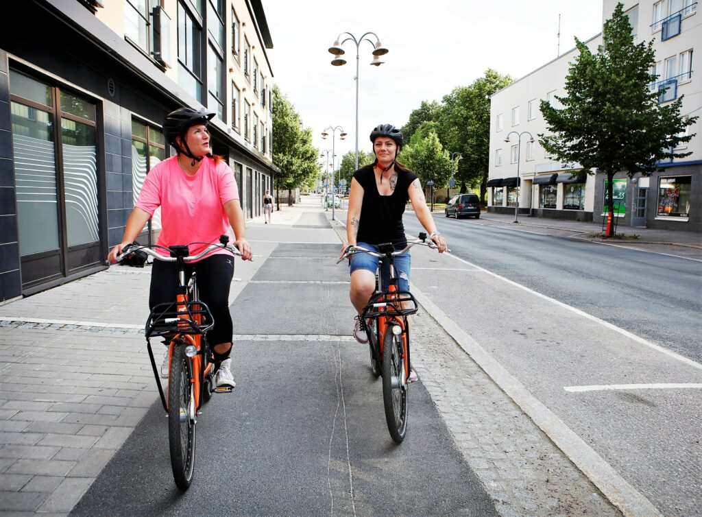 Kaksi naista pyöräilee kadulla Riihimäen keskustassa. On kesä. Pyörät ovat oransseja kaupunkipyöriä.