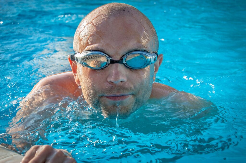 Kalju mies uimassa musta reunaiset uimalasit päässä. Kuva otettu suoraan edestä päin vesirajan tasolta.