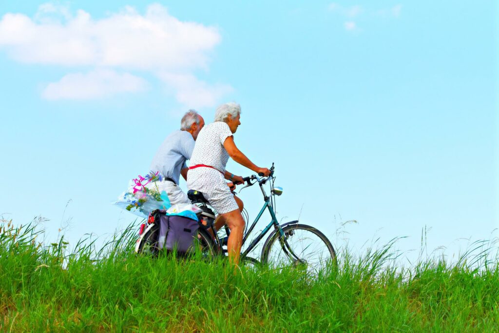 Kaksi vanhempaa henkilöä pyöräilevät kesällä vehreässä maastossa.