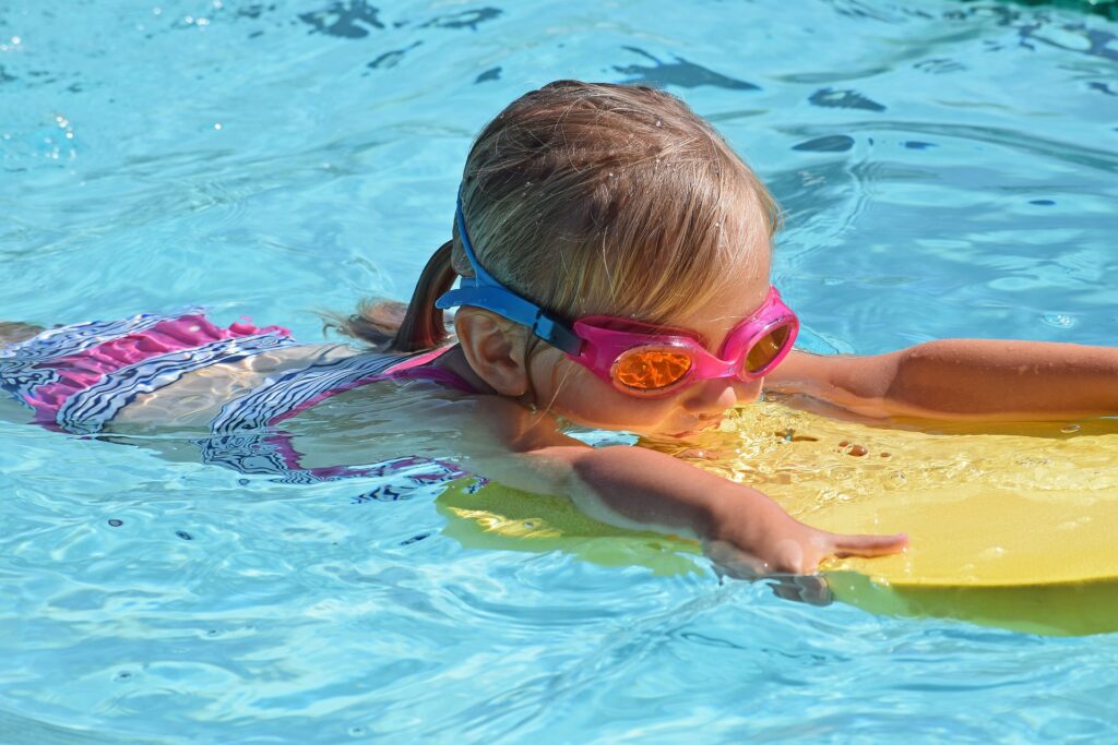 A child swims on a swim board in a swimming pool. The child has pink bathing suits and a yellow swimming board.