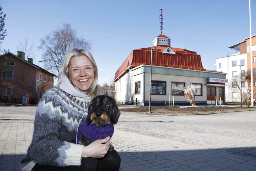 Hymyilevä Sonja Kailassaari, jolla on sylissä mäyräkoira. Taustalla Kino Sampo -rakennus ja sininen taivas.