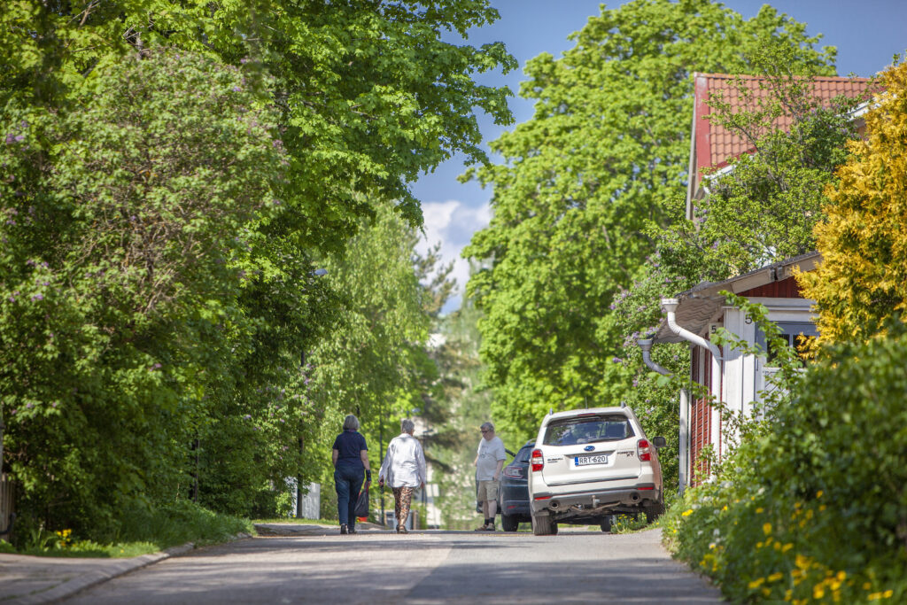 Maisemakuva Riihimäen Maantiestä. Kesä. Tiellä näkyy osittain puutalo ja teillä myös auto ja kolme ihmistä.