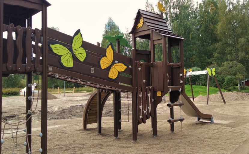 Hiekkakentällä kookas puinen lasten kiipeilyteline, jossa suuria perhosia. Lehtipuita taustalla.
