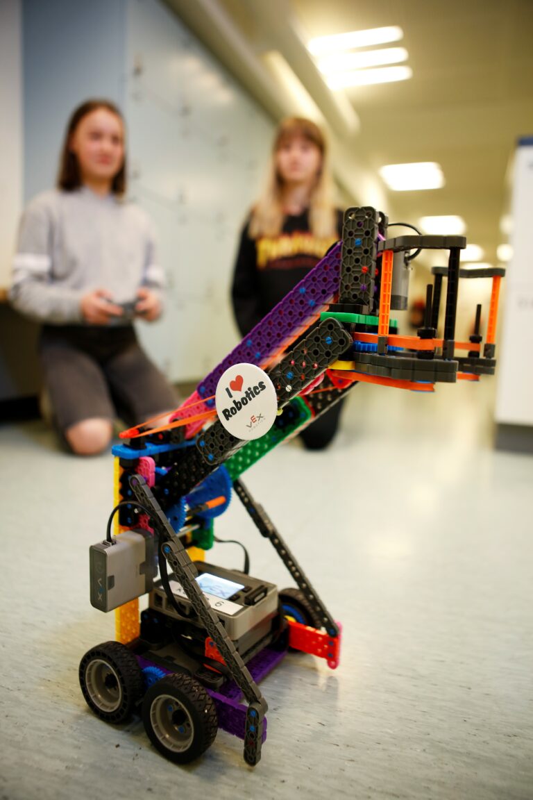 A robot in the foreground and two teenage girls blurred in the background.