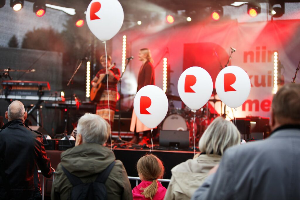 Riihimäki-päivän keikkakuva, jossa näkyy valkoisia R-ilmapalloja, esiintyjiä lavalla ja yleisöä lavan edessä.