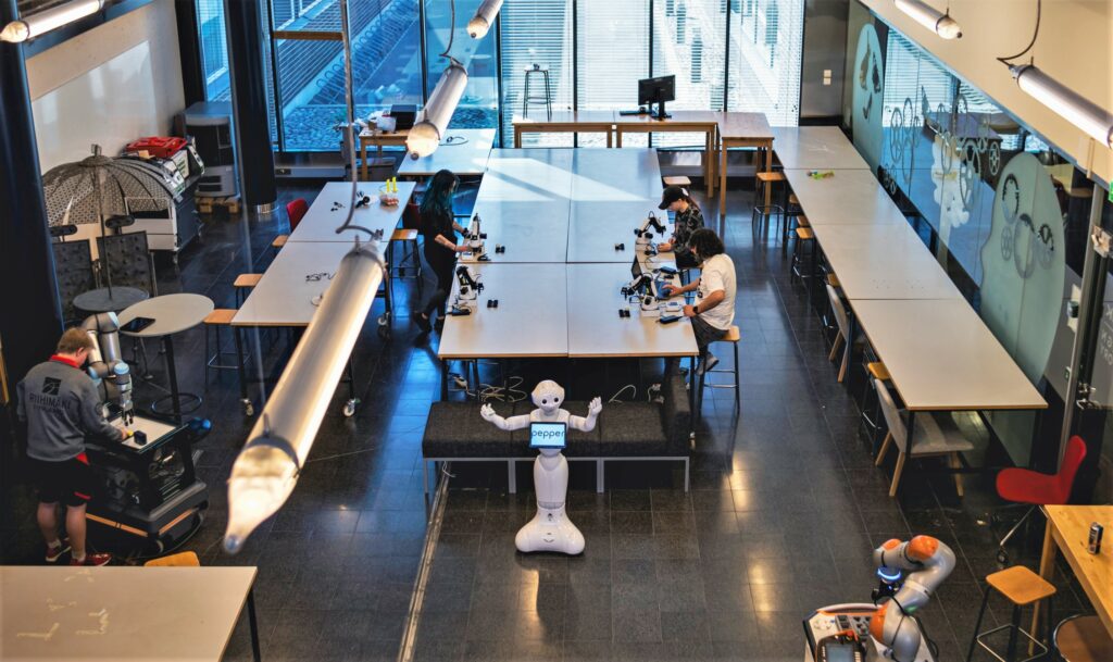 Yleiskuva RoboLabin luokasta, jossa ihmisiä, suuri työskentelypöytiä ja robotteja.