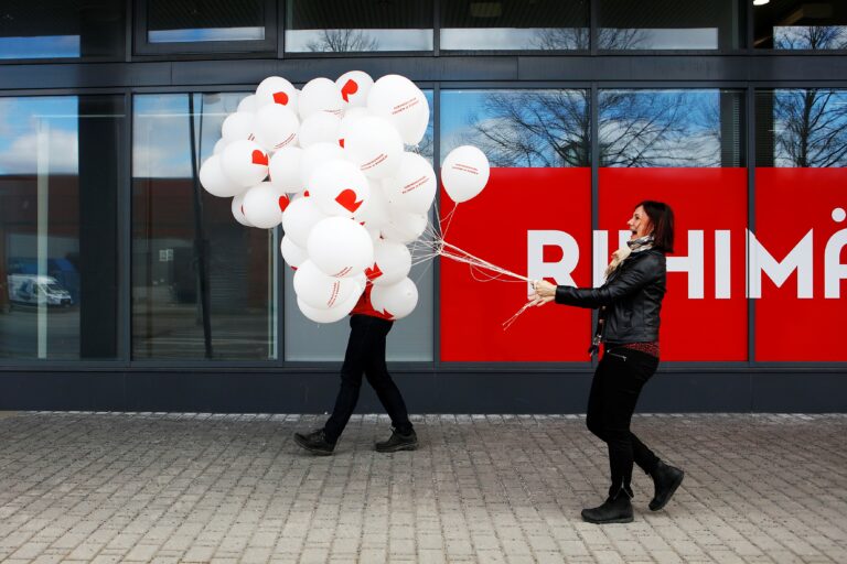 A woman is walking down the street with a big bunch of white R balloons in her hand. Another person's legs are visible from behind the ball bundle and a partial Riihimäki logo is visible in the window.