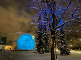 Snowy picture of dark time in Kirjastonpuisto's experience museum. The tree in the foreground is lit with blue light and the text is projected on the wall of the library