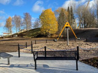 Aurinkoisen syksyllä otetun kuvan etualalla kaksi mustaa penkkiä, joiden takana vauhdikas karuselli kahdelle. Kuvan takaosassa mäki, jossa kuntoportaat.