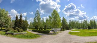 Aurinkoinen kesäkuva Tehtaankadun messupuistosta, missä näkyy puita, nurmialuetta, hiekkateitä ja pieni kaarisilta.