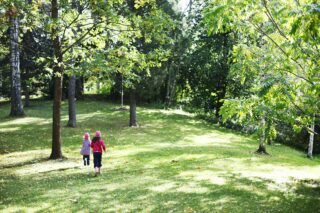 Kaksi lasta juoksee kesällä Rautatienpuiston nurmella puiden siimeksessä. auringon paistaessa.