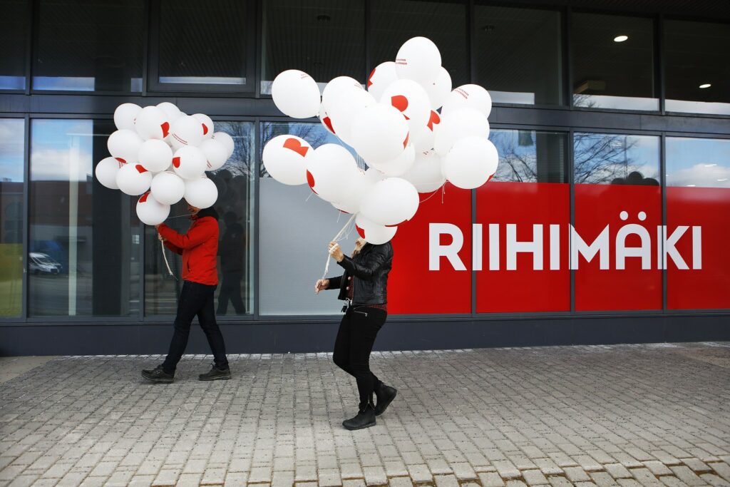 Two people walking with bunches of balloons. Face covered in balloons. Behind the wall with the logo of the city of Riihimäki.