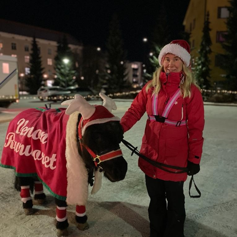 Nainen ja poni pimeässä illassa. Naisella tonttulakki ja ponilla poronsarvet ja punainen viitta.