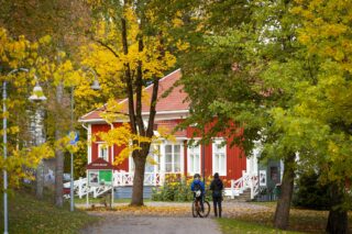 The red wooden house of the city museum in the Railway Park in autumn. In the picture, two young people are standing in front of the museum in the shade of big oak trees.