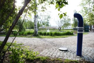 A sound piece in Jukka Jalonen's park, which includes a tile on the ground and a blue and white pipe. A pond and a fountain can be seen in the background.