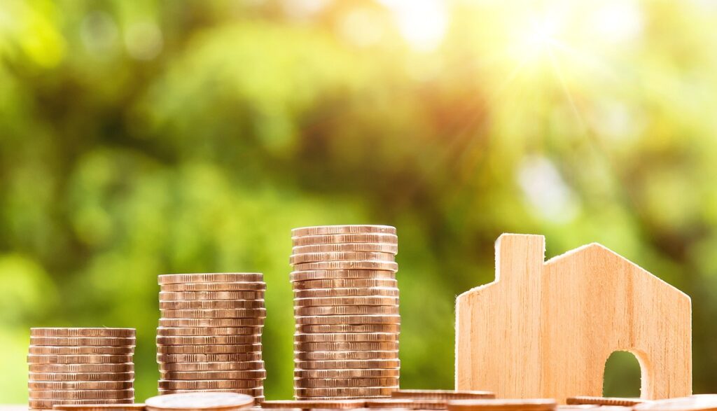 Three stacks of coins with a small wooden house next to them. Blurred tree branches in the background.