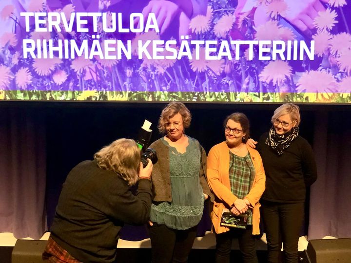 Kolme naista valokuvattavana. Taustalla violetti kangas jossa teksti Tervetuloa Riihimäen kesäteatteriin. Etualalla valokuvaaja selin kameraan.