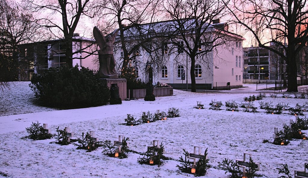 Talvinen näkymä sankarihautausmaalle. Etualalla sankarihaudat ja taustalla enkelipatsas ja valkoinen talo.