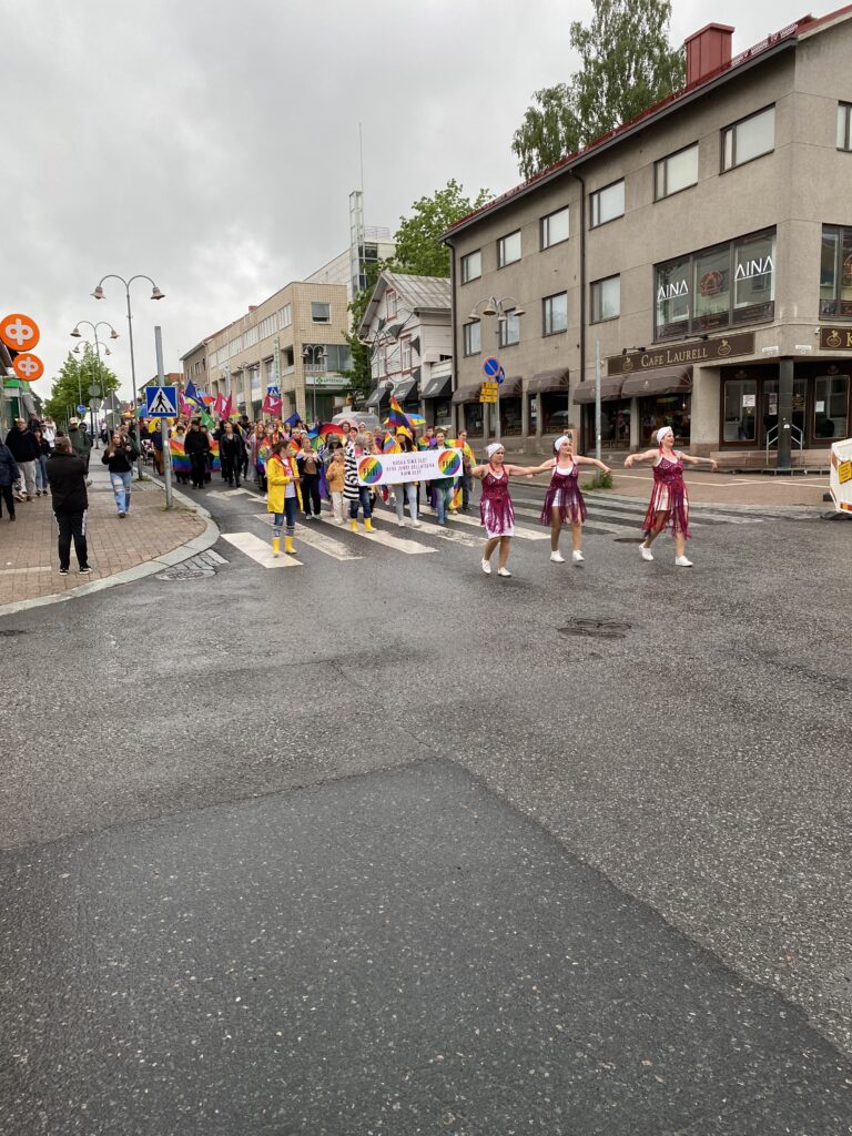 There is a pride parade with dancers along the rainy street.