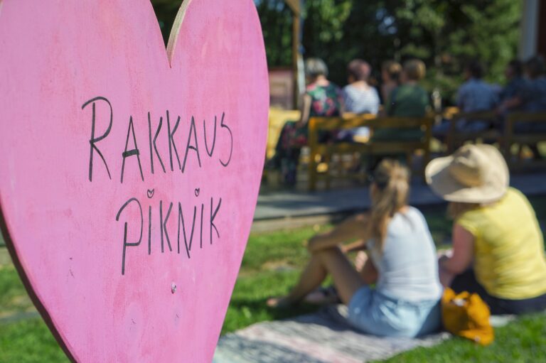 Vaaleanpunainen sydänkyltti, jossa lukee Rakkaus-piknik. Takana istuu ihmisiä maassa.