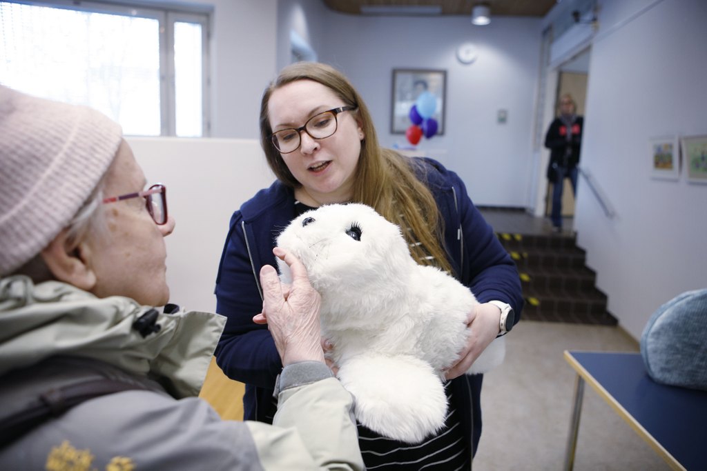 Iäkäs nainen silittää valkoista robottihyljettä, jota nuorempi nainen pitää sylissään.