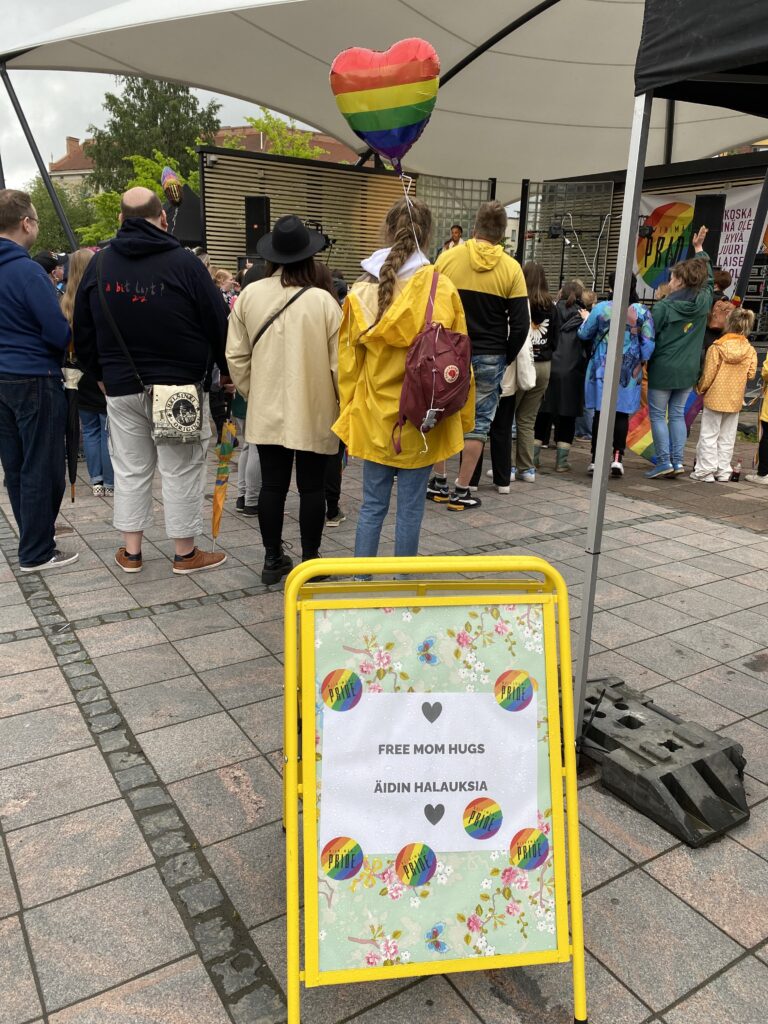 In front, a yellow vertical stand with the words "mothers' hug point". Behind are people and a pride balloon.