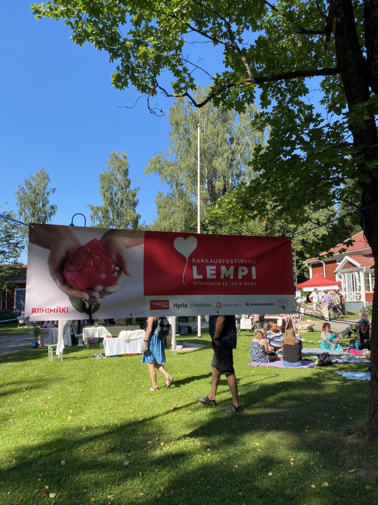 Banderolli, jossa lukee Lempi. Banderollin takana näkyy vihreä piha ja ihmisiä.