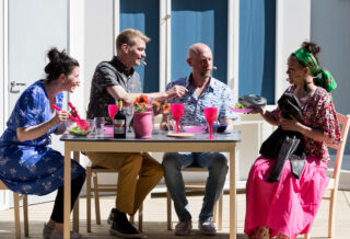 neljä henkilöä ruokapöydän ääressä ruokailutilanteessa kesäteatterilavalla