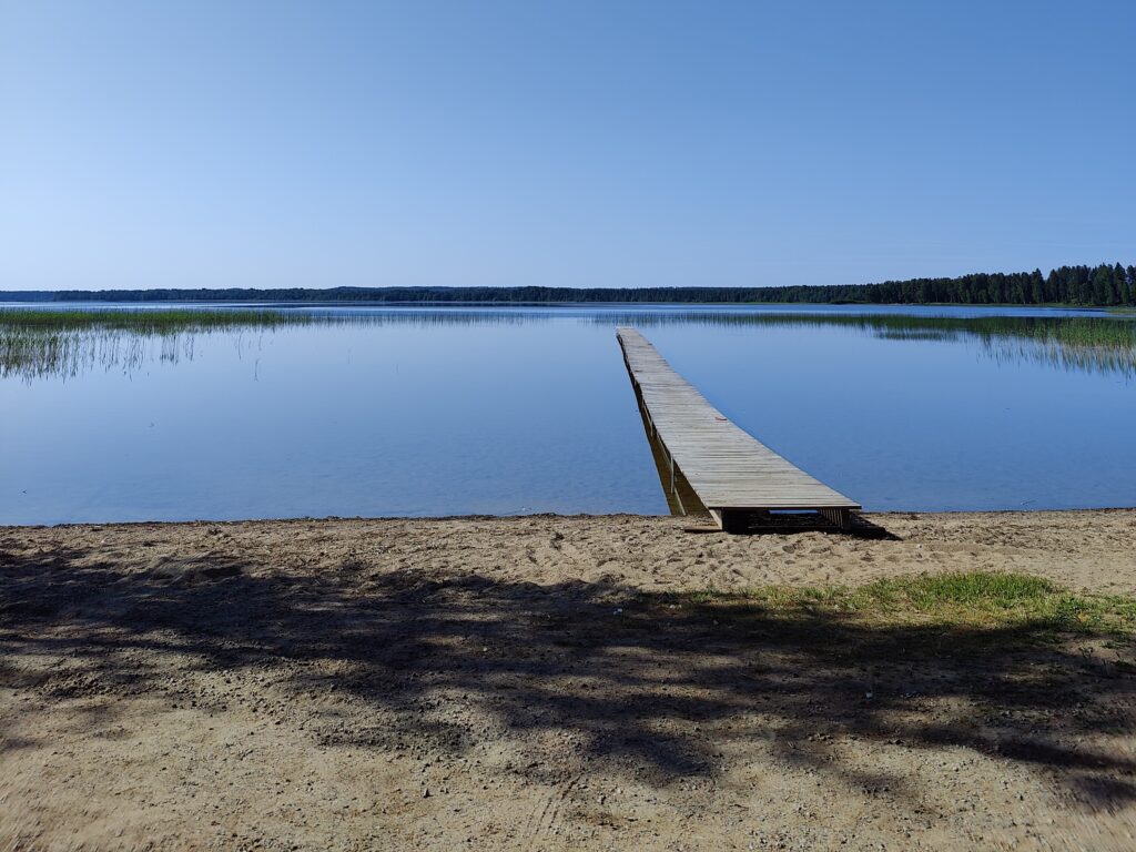Kesäinen uimaranta, jossa ei ole ihmisiä, pitkä laituri tyyneen veteen.