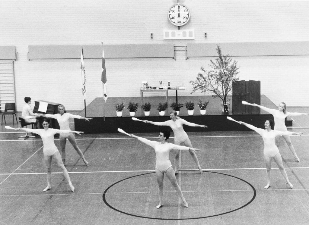 Mustavalkoinen valokuva vuodelta 1970, jossa kuusi naista voimistelee liikuntasalissa pianon säestäessä.