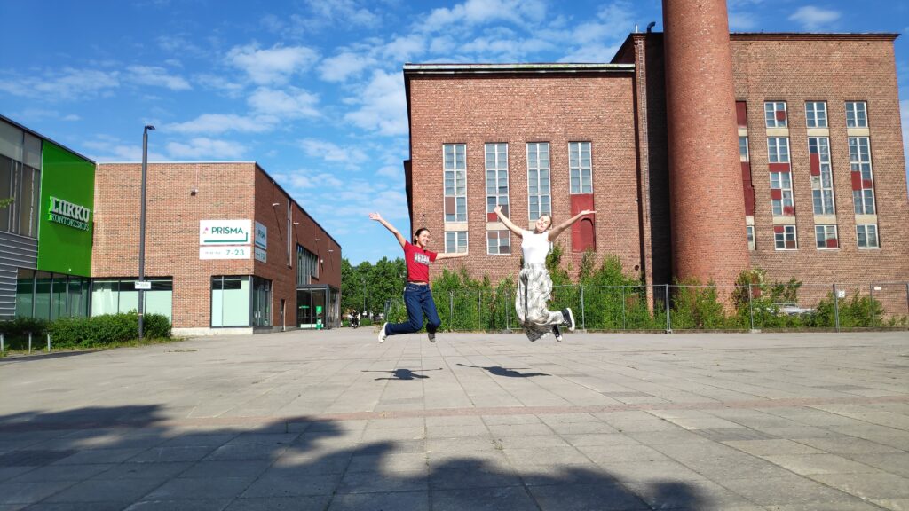 Kaksi nuorta hyppäämässä ilmaan aukiolla Riihimäen Voimala-rakennuksen edessä kesäisessä säässä.