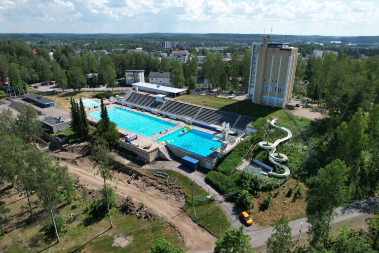 In the summer sun, the in-ground swimming pool shimmers turquoise, next to it is a high water tower and a swimming hall, next to which is a gravel road along which construction site vehicles travel.