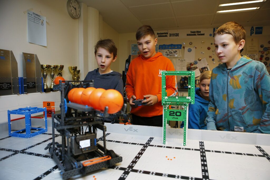 Kolme lasta hämmästelee oransseja palloja kantavaa robottia.