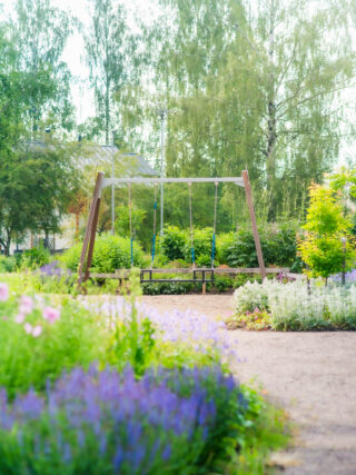 The park has car seat swings for small children where parents can also swing. The swings are surrounded by vegetated areas with lots of blue-tinged flowers.