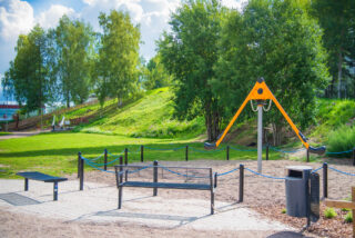 In the foreground of the photo, taken on a sunny summer day, there are two black benches, behind which is a speedy carousel for two. In the back of the picture, a hill with fitness stairs.