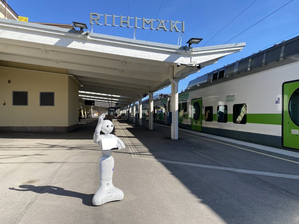 Pepper-robotti seisoo vilkuttamassa asemalaiturilla. Taustalla asemarakennus, jossa lukee Riihimäki. Taustaalla myös laiturin viereen pysähtynyt juna.