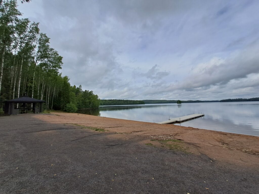 Kesäinen uimaranta tyhjillään. Rannassa on laituri ja vesi rajoittuu puustoon.