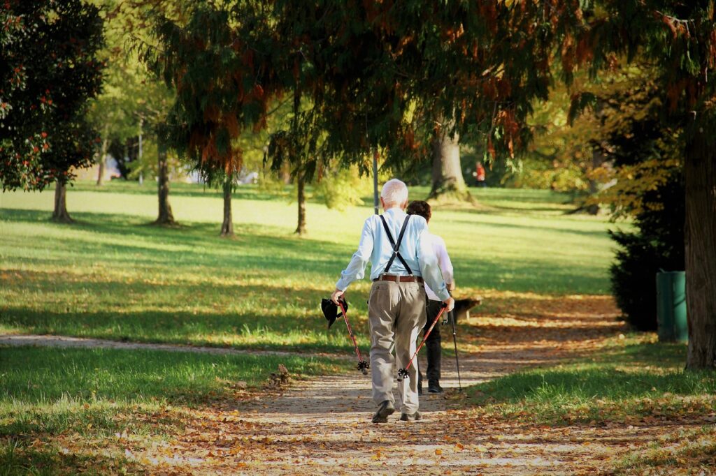 Kaksi vanhempaa henkilöä ulkoilemassa keväisessä puistossa, kävelemässä poispäin kamerasta.