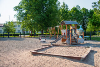 The park's fenced playground has many different play equipment. In the foreground is a sandbox and a train-themed slide.