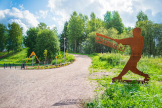 In the foreground is a metal statue holding a Piikinmäki sign. The carousel and trees in the playground can be seen in the background.