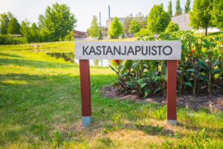 Kastanpuisto's wooden sign, behind is a planting area and a pond.