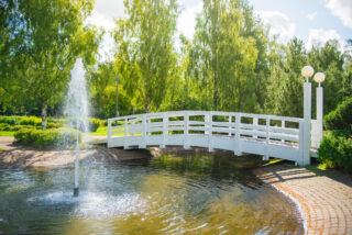 There is a small water feature in Messupuisto over which a wooden arched bridge goes. The water feature also has a fountain, surrounded by vegetation.