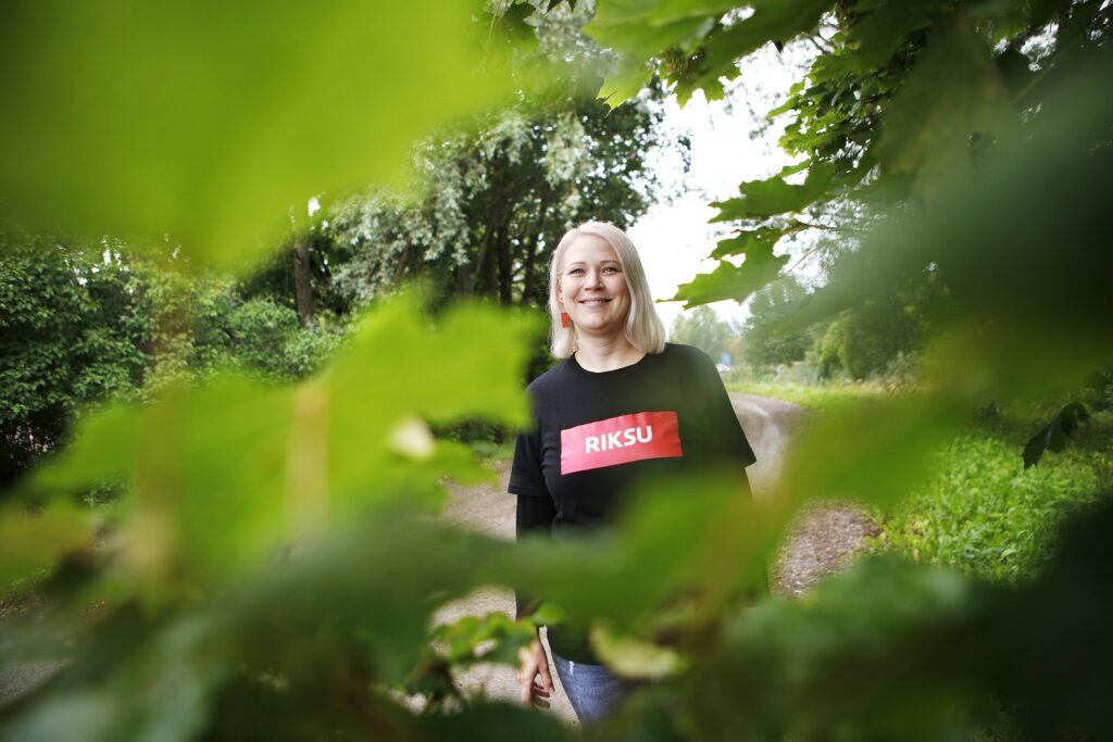 Between the green leaves is a smiling person whose black shirt reads Riksu.