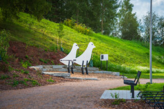 Gritbird exercise equipment with plants around it and an instruction board for use during the exercise interval. In the foreground of the picture is a black bench and a bike rack.