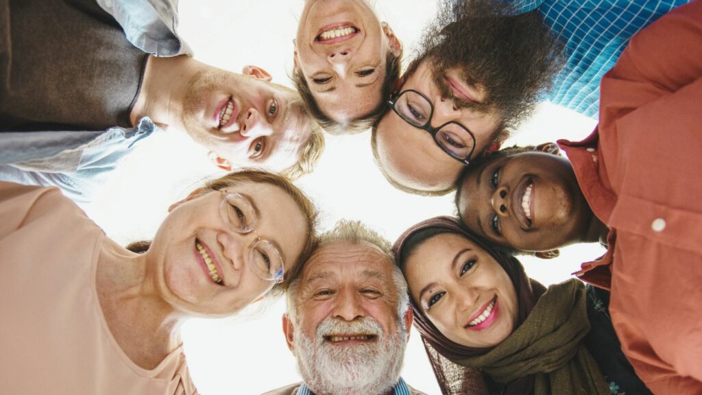 The smiling faces of seven people of different looks and ages in a circle, shot from below.