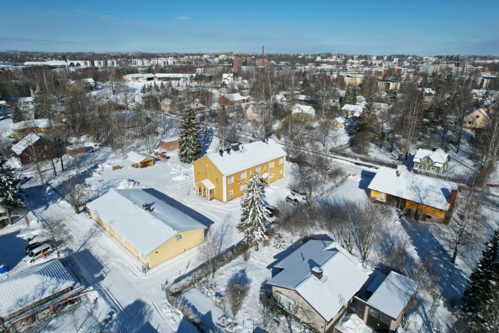 Viistoilmakuva Patastenmäen puukoulun alueesta. Kuvan keskellä on keltainen, kaksikerroksinen, puukoulu. Kuvassa näkyy ympäröivää rakennuskantaa. Kuva on otettu aurinkoisena talvipäivänä.