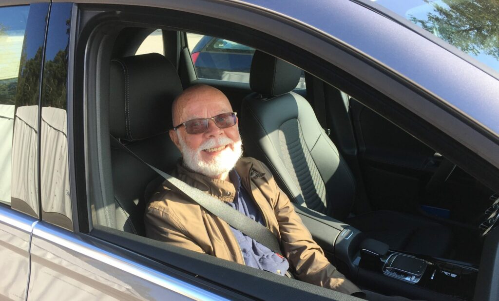 Autossa etupenkillä istuva vanhus katsoo hymyillen avoinaisesta auton ikkunasta ulos aurinkoisessa säässä.