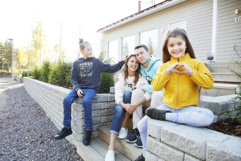 A girl sits on a low wall in front of a pale detached house, her hands forming a heart symbol. The mother and father of the family are sitting next to him on the stairs. In the background, a boy is sitting on the wall, one of his hands is in his mother's hair.